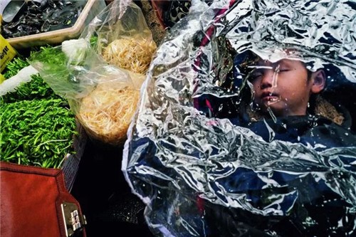 在紐約唐人街，你能買到最新鮮的魚、肉和蔬菜，也能吃到最正宗的中國菜，圖中，菜市場(chǎng)的孩子在菜攤旁熟睡。