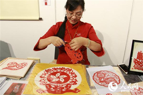 潮陽剪紙傳承人魏惠君現(xiàn)場展示，她的面前就擺放著專為此次展覽設(shè)計(jì)的“20”(左一)和區(qū)徽(左二)圖樣。(攝影：吳玉潔)